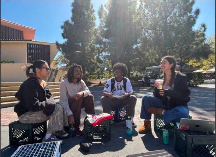 Four students sitting on milk crates outside the studio, talking